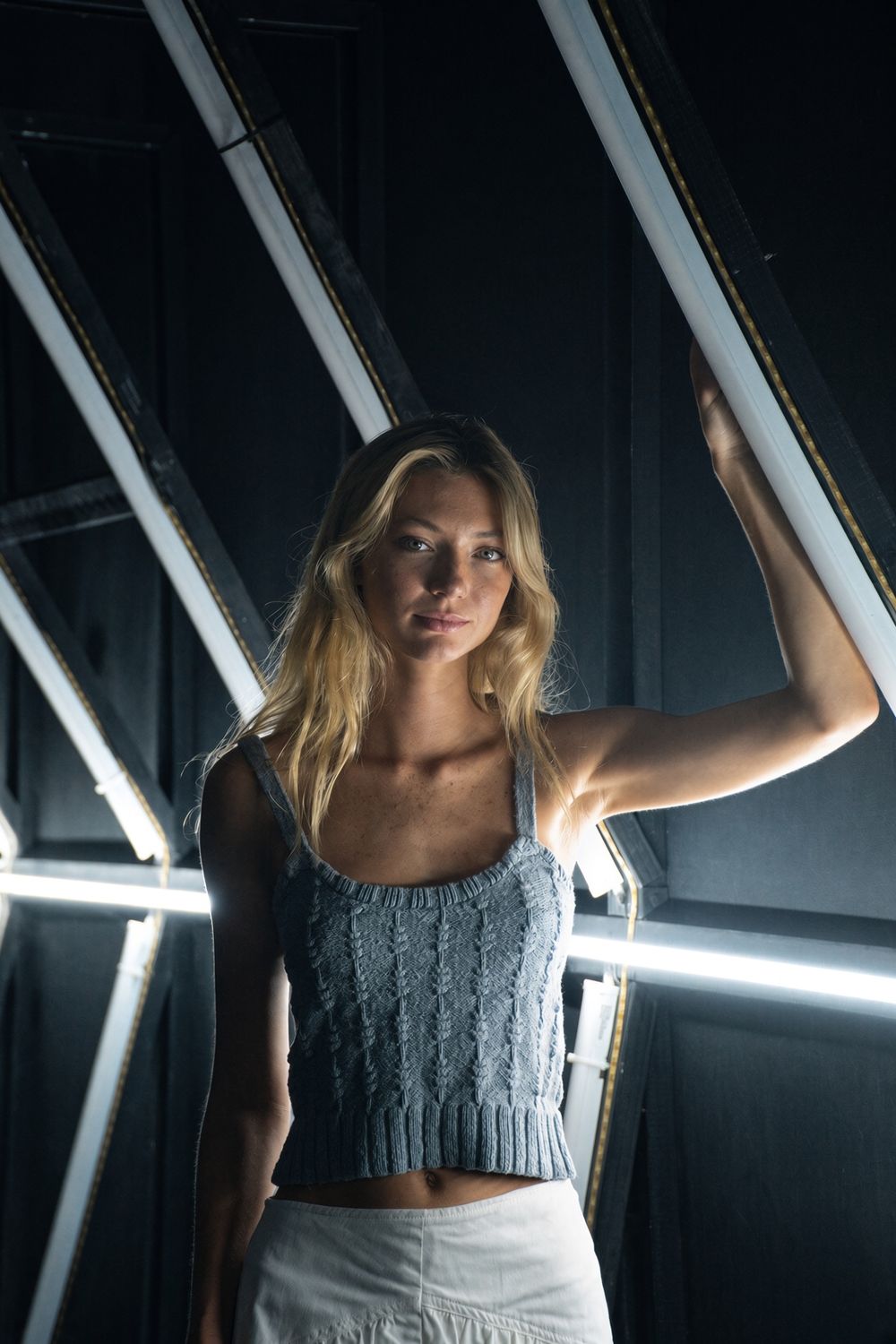 A woman in a blue knit top raises one hand to a glowing light tunnel in a dark studio.