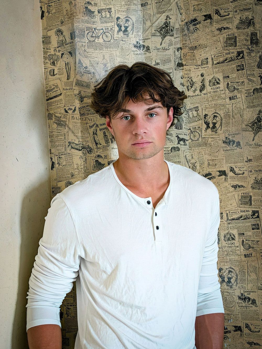 A man in a white long-sleeved henley stands in front of vintage newspaper-print wallpaper.