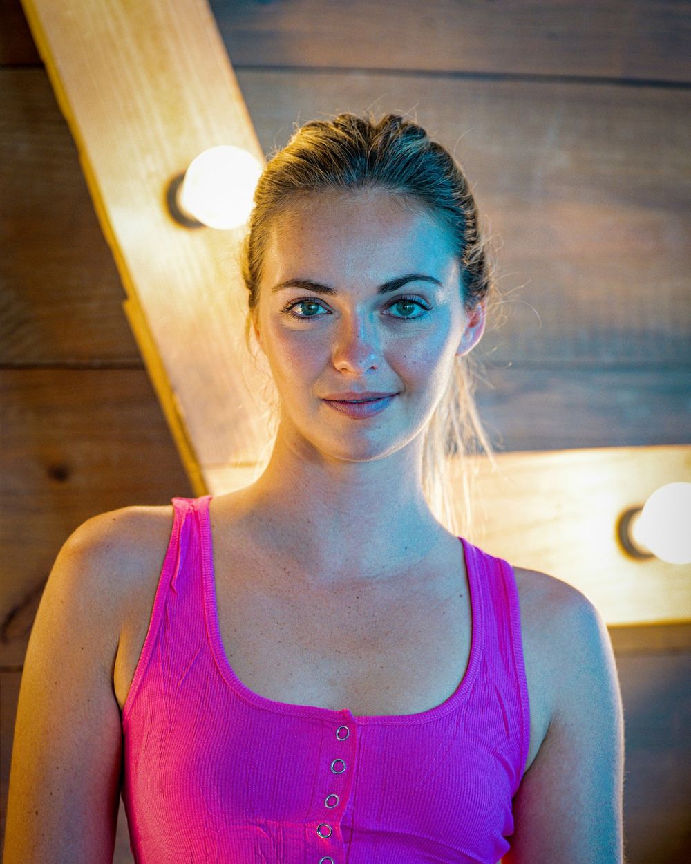 A woman in a magenta tank top is lit by warm marquee bulbs against a wooden wall.