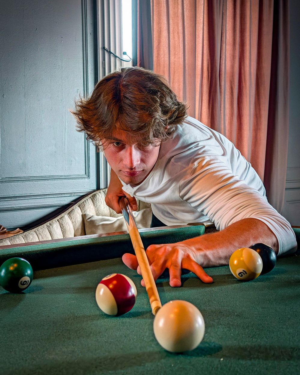 A man in a white henley lines up a shot at a pool table in a room with tall windows and rose curtains.