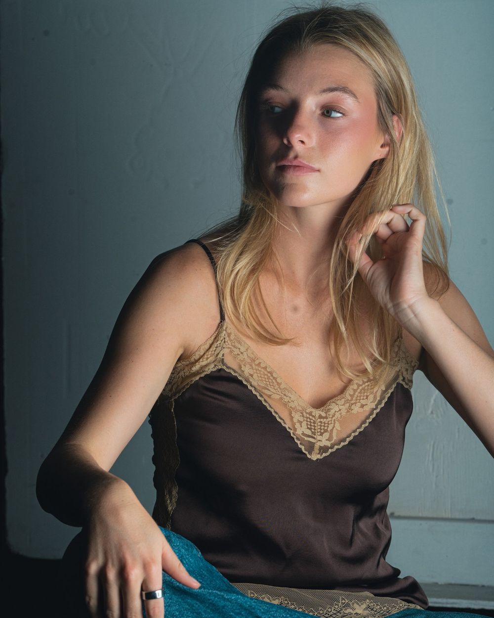 A woman in a brown satin camisole with lace trim sits against a dark teal backdrop, one hand at her neck.