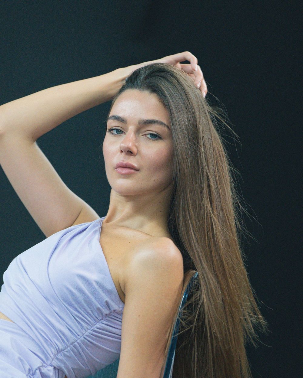 A woman with long dark hair in a lilac one-shoulder top lifts one arm against a dark teal backdrop.