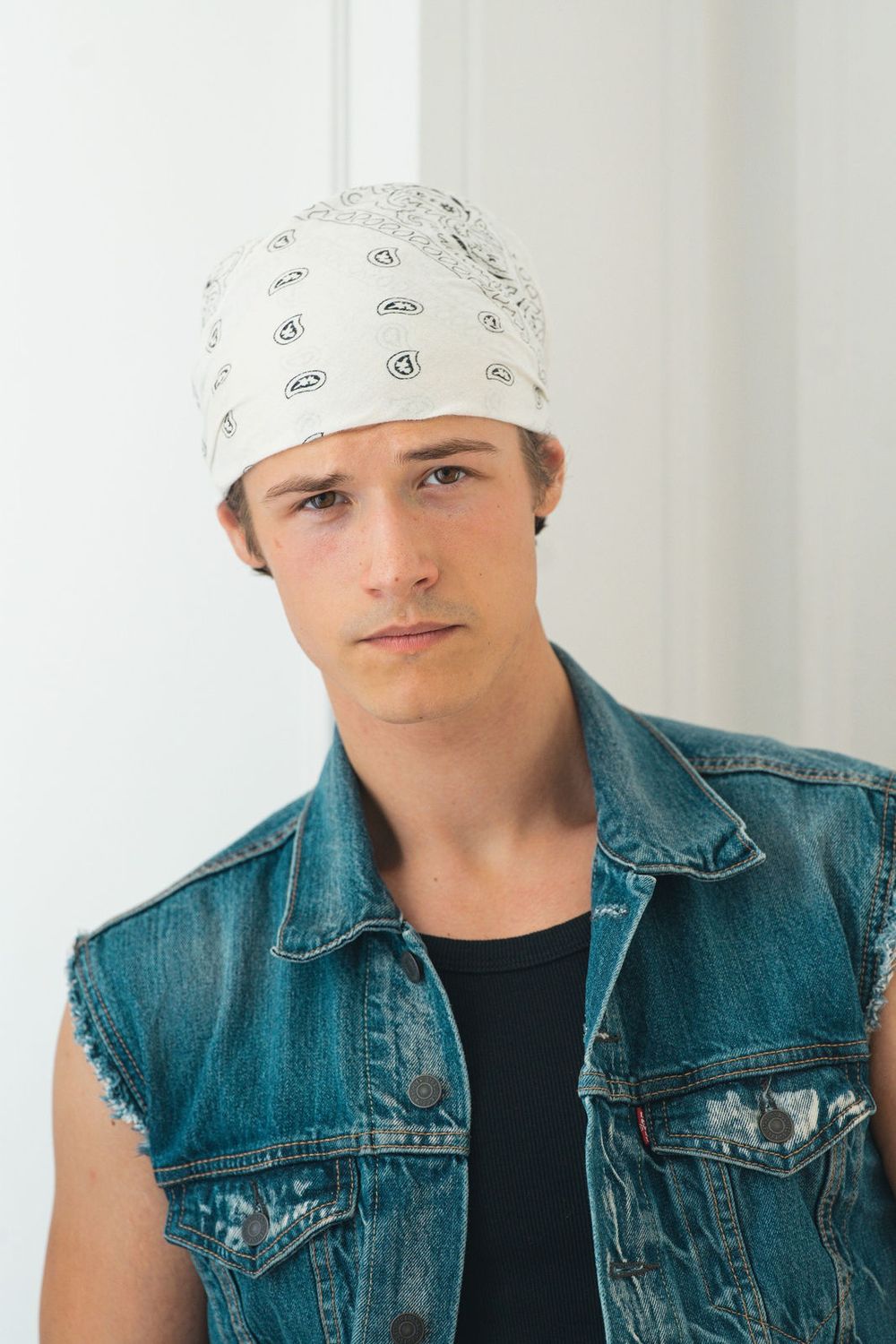 A man in a sleeveless denim vest wears a white bandana tied over his hair.