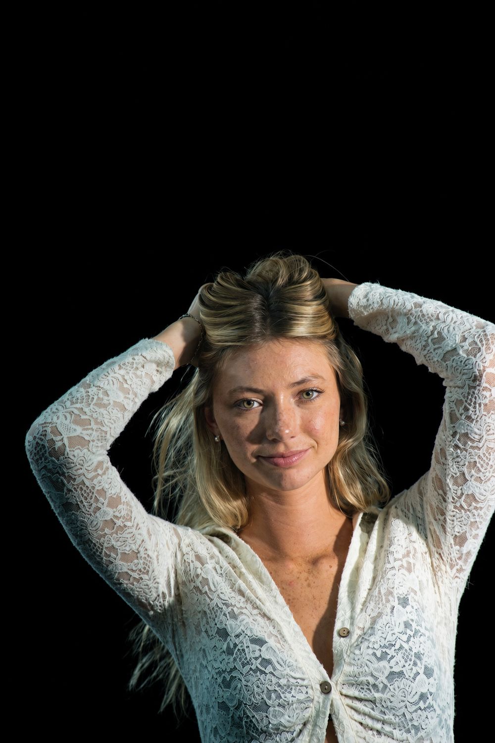 A smiling woman in a cream lace cardigan holds her hair up with both hands against a black backdrop.