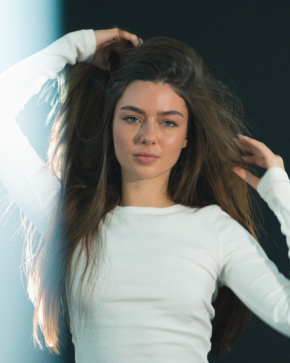A woman in a white long-sleeved top runs her hands through long dark hair in soft window light.