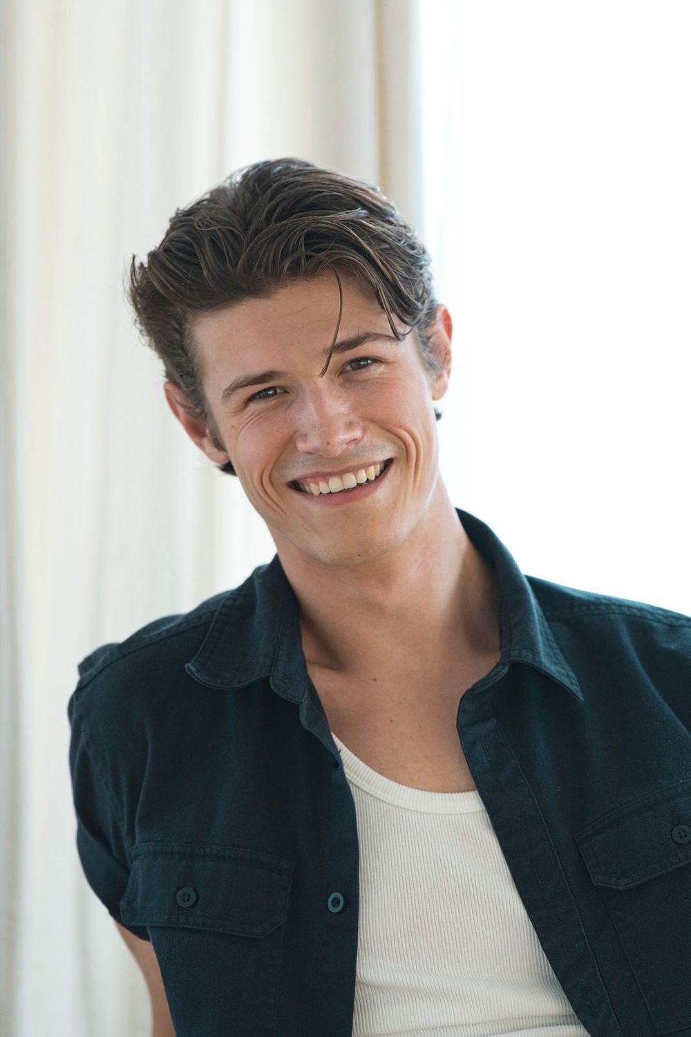 A man in a dark teal short-sleeve shirt worn open over a white tank top smiles in a bright room with cream curtains.