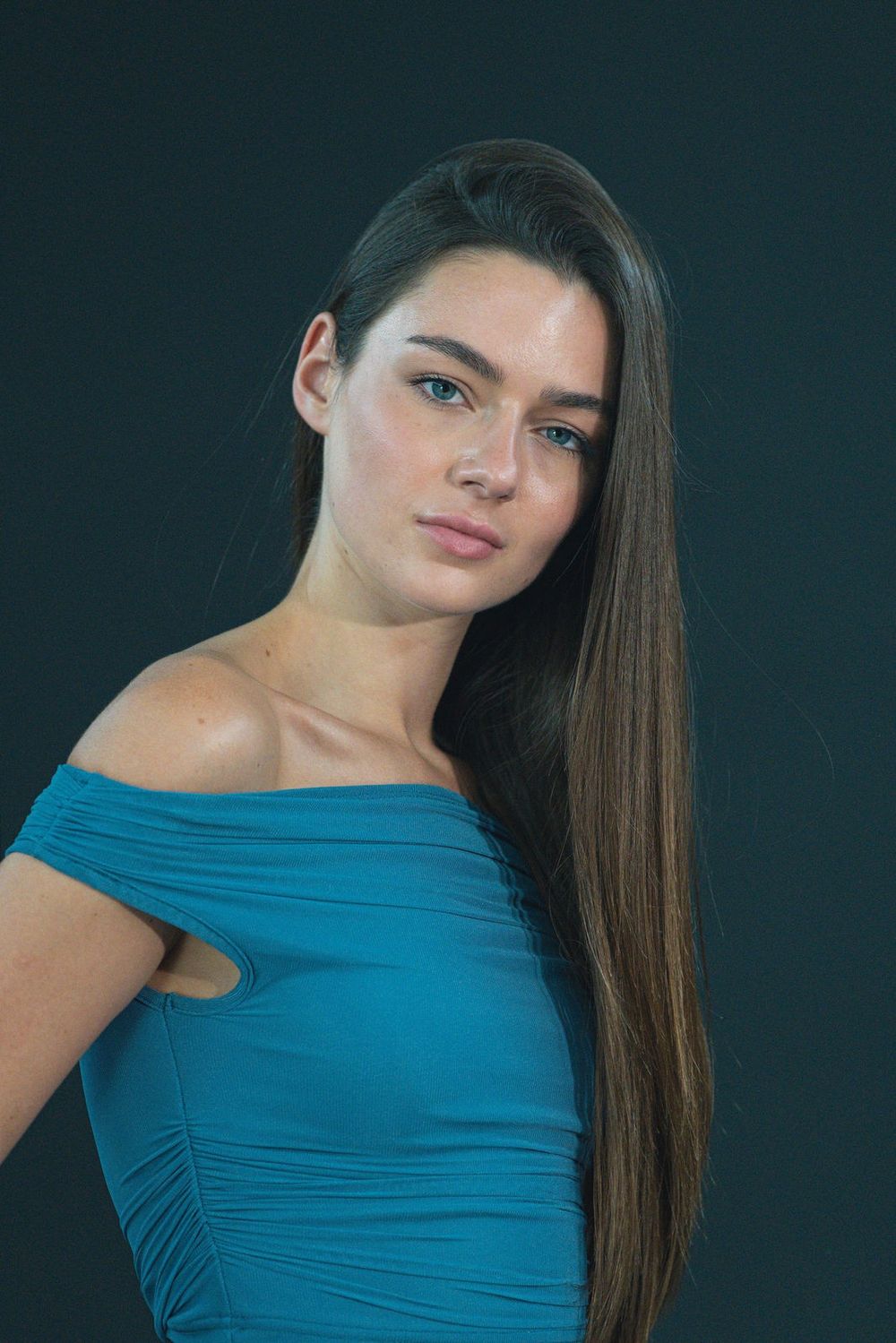 A woman with long dark hair in a teal off-the-shoulder top tilts her head in front of a dark teal backdrop.