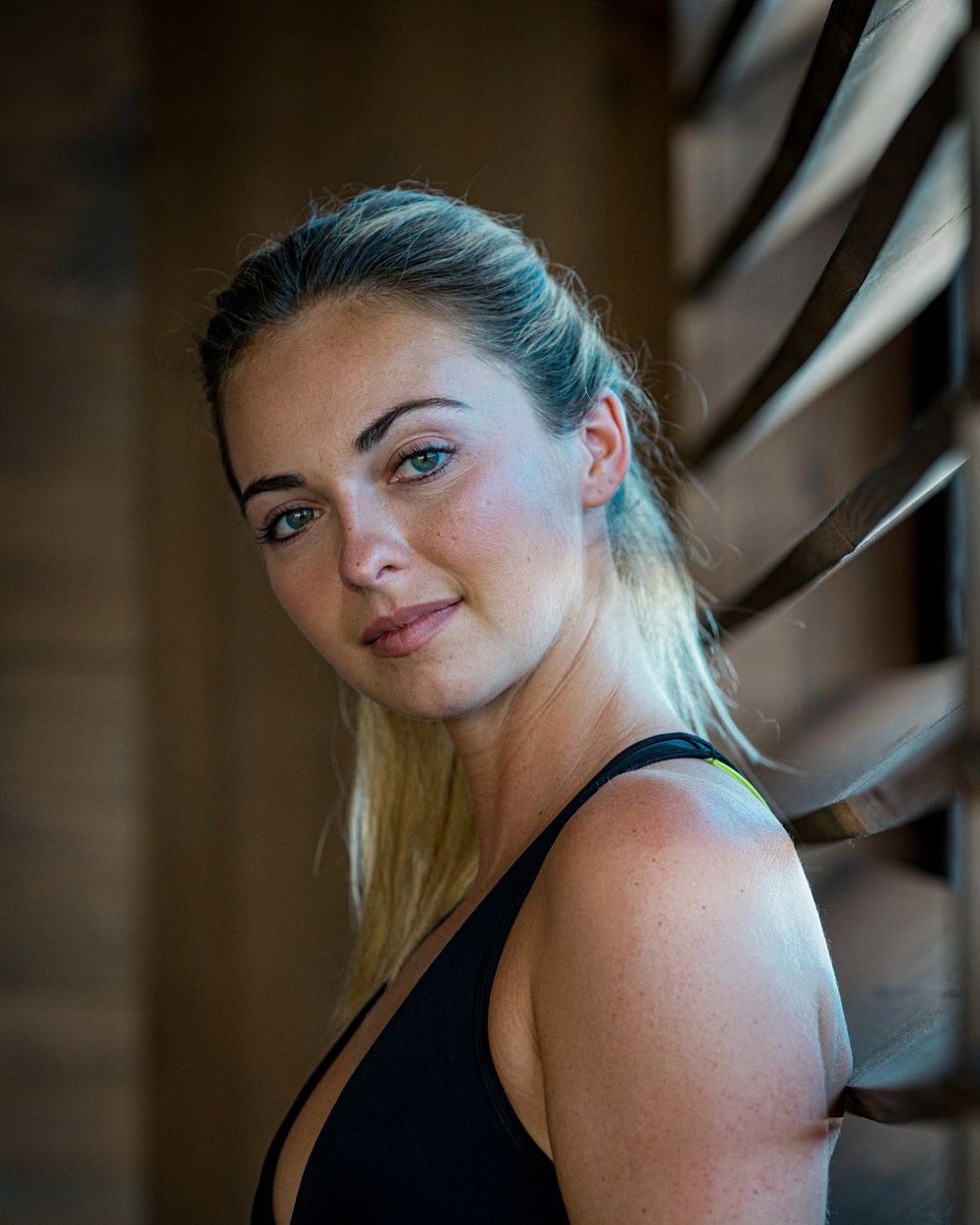 A woman in a black top is lit from the side in front of dark wooden slats.