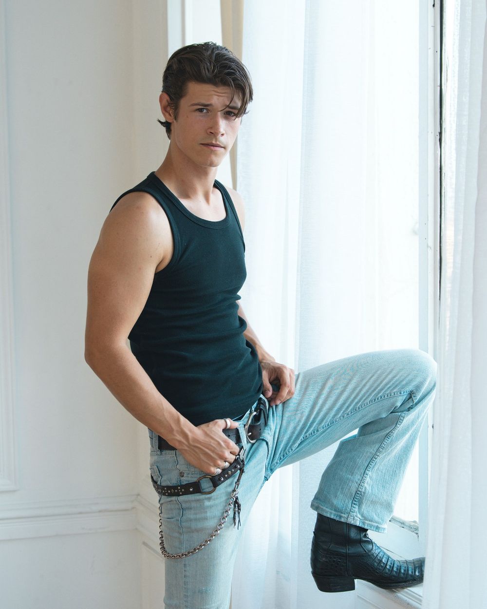 A man in a black tank top and light jeans rests one boot on a white window ledge.