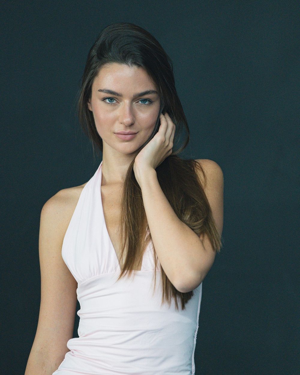 A woman in a white halter top rests her cheek on her hand against a dark teal backdrop.