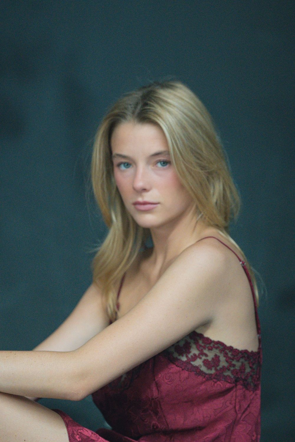 A woman in a wine-coloured satin slip dress with lace trim sits on a glossy floor in front of a dark teal backdrop.