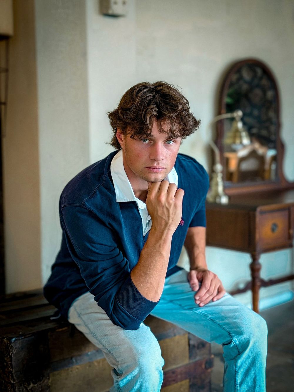 A man in a navy rugby shirt and light blue jeans sits on an old trunk with his chin on his hand, a desk and an arched mirror behind him.