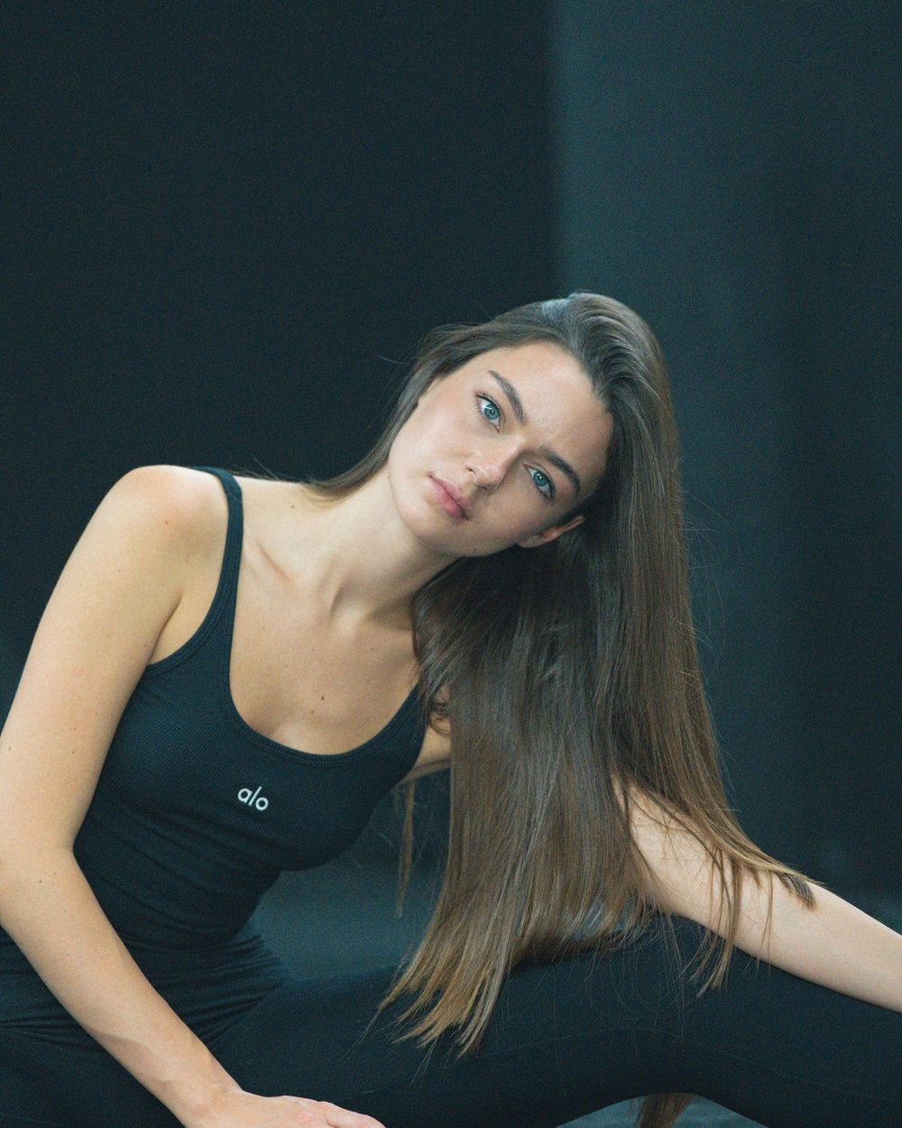A woman in a black tank top leans to one side, her long dark hair falling over her shoulder.