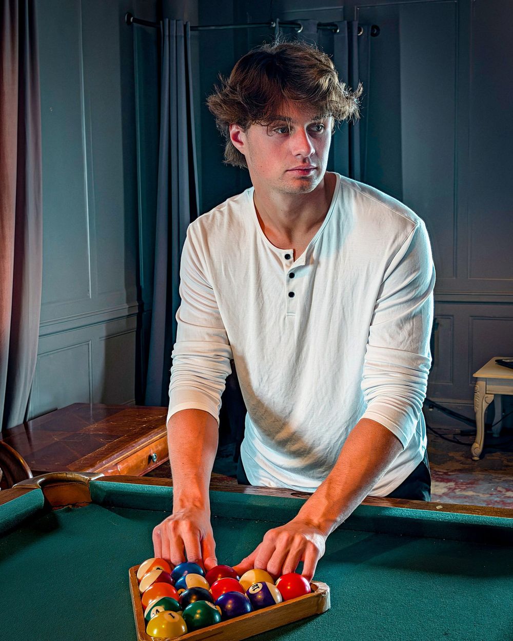 A man in a white henley racks pool balls on a green table.