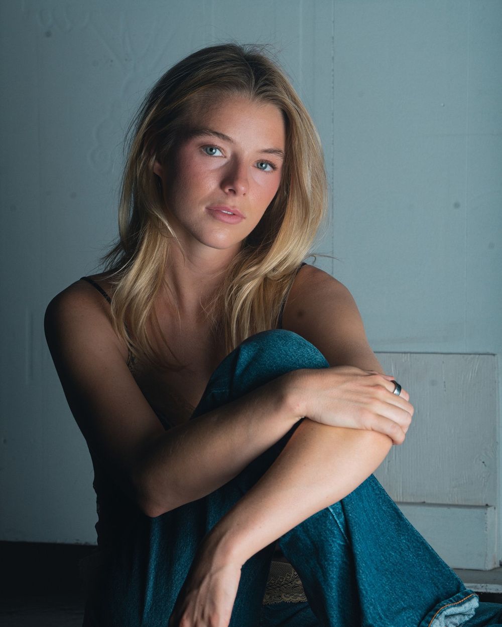 A woman in jeans sits with her arms around one knee against a grey-blue wall.