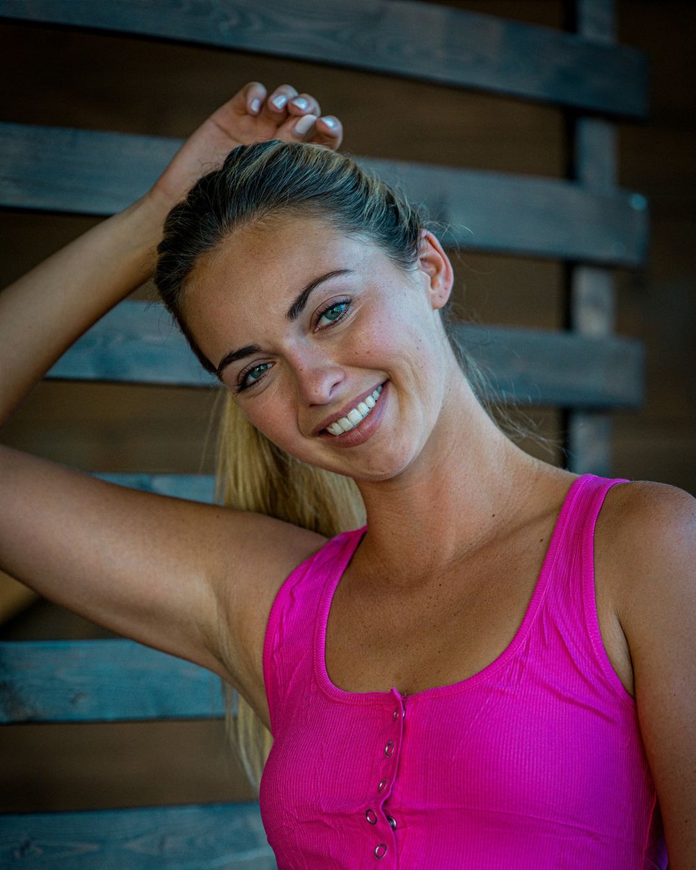 A smiling woman in a pink tank top lifts one arm against a wooden wall.