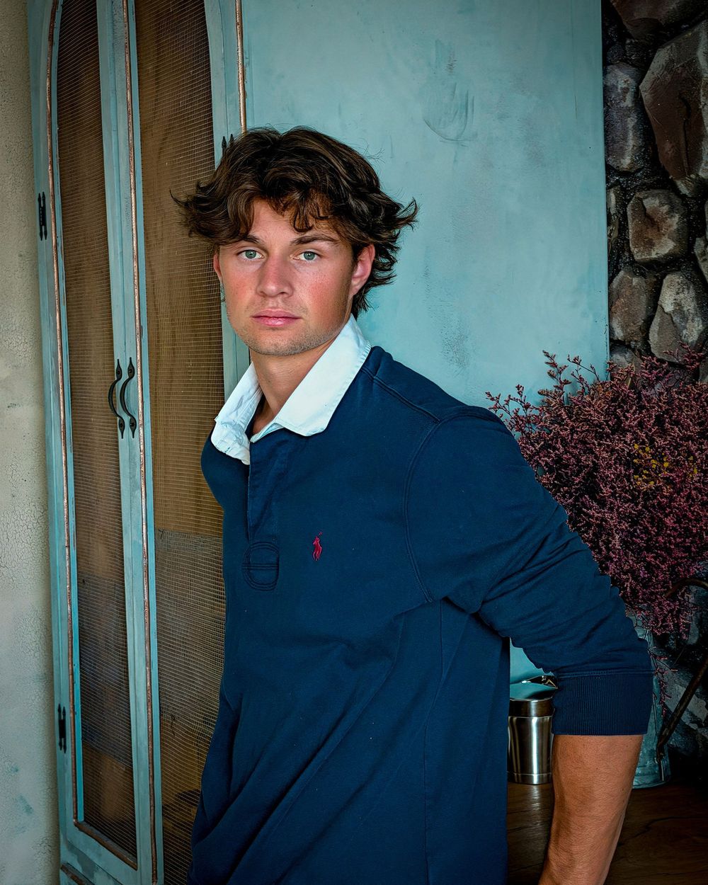 A man in a navy polo leans in a doorway beside a stone fireplace and dried flowers.